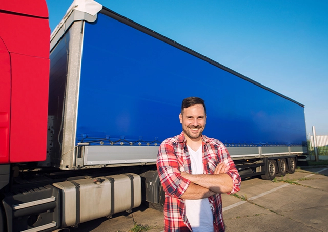 Smiling man in a red plaid shirt stands with arms crossed in front of a large blue delivery trailer attached to a red truck.