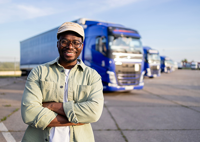 A smiling truck driver with arms crossed standing on parking lot.