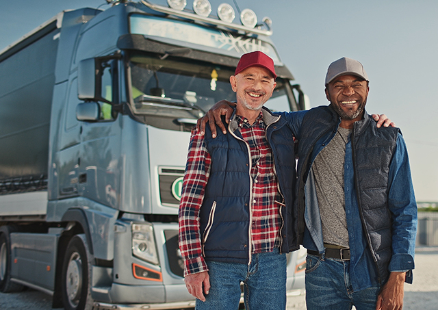 Two professional, multicultural truck drivers smiling and looking at the camera, with their vehicles parked behind them.